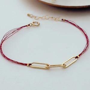 Könnte beinhalten: Ein rotes Armband mit einer goldenen Kette und drei goldenen ovalen Gliedern.