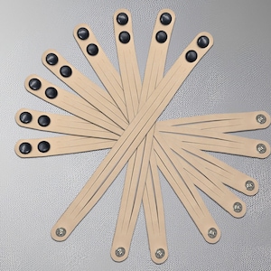 May include: A collection of light tan leather wristbands, each with black snap closures and silver rivets. The wristbands are arranged in a circular pattern, showcasing their design and potential for use as accessories.
