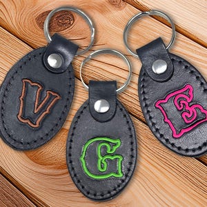 May include: Three black leather keychains with silver key rings. Each keychain has a different initial: a brown "V", a green "G", and a pink "E". The initials are stitched onto the oval-shaped keychains.