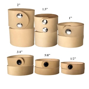 May include: A selection of light tan leather cuff bracelets in various sizes, ranging from 1.3 cm to 5.1 cm wide. Each cuff features a snap closure with either silver or black hardware. The cuffs are stacked to show the different sizes.