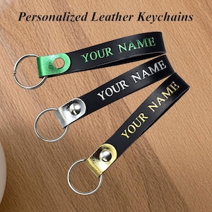May include: Three black leather keychains with silver key rings. Each keychain is personalized with the text "YOUR NAME" in green, silver, and gold. The text "Personalized Leather Keychains" is at the top.
