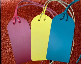 May include: Three leather luggage tags in shades of purple, yellow, and teal. Each tag has a matching leather loop for attachment. The tags are displayed against a textured, reddish-brown background.