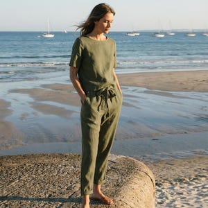 May include: An olive green linen two-piece outfit, consisting of a short-sleeved top and cropped pants with a drawstring waist. The outfit is worn with brown sandals. The background features a beach, ocean, and sailboats.
