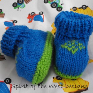Op de afbeelding: Een paar blauwe en groene gebreide babyschoentjes met een groen en blauw ontwerp op de bovenkant. De schoentjes liggen op een witte achtergrond met een patroon van speelgoedtrucks.