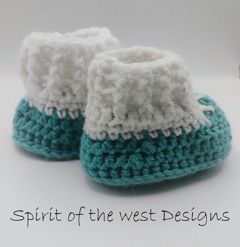 Pu&ograve; includere: Un paio di scarpine da neonato all'uncinetto fatte a mano. Le scarpine hanno una base turchese e un polsino bianco soffice. Il testo "Spirit of the west Designs" &egrave; scritto in corsivo nero nella parte inferiore dell'immagine.