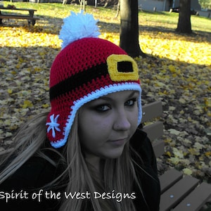 May include: A red and white crocheted hat with a black band and a yellow buckle. The hat has a white pom-pom on top and a snowflake design on the side. "Spirit of the West Designs" is written on the bottom of the image.