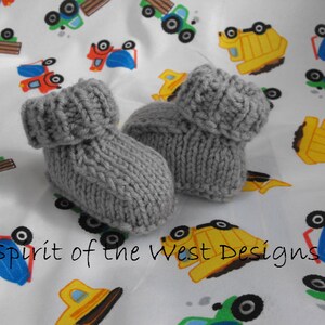 Op de afbeelding: Een paar grijze gebreide babyschoentjes met een omslagboord. De schoentjes liggen op een witte achtergrond met een patroon van kleurrijke speelgoedtrucks.