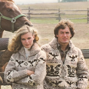 May include: A couple wearing matching brown, white and black patterned knit sweaters. The sweaters feature a geometric design with birds and trees. The man is standing with his hands in his pockets, and the woman is standing with her arms crossed. They are standing in front of a wooden fence and a brown horse.