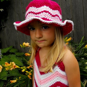 May include: A handmade crochet hat and dress set in shades of pink and white. The hat features a wavy pattern and a white scalloped brim. The dress has a chevron design. The text "Spirit of the West Designs" is visible.