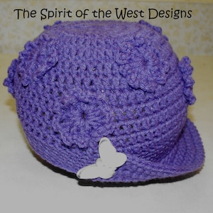 May include: Purple crocheted hat with a white butterfly embellishment. The hat has a brim and a textured design. The Spirit of the West Designs is written in the top left corner of the image.