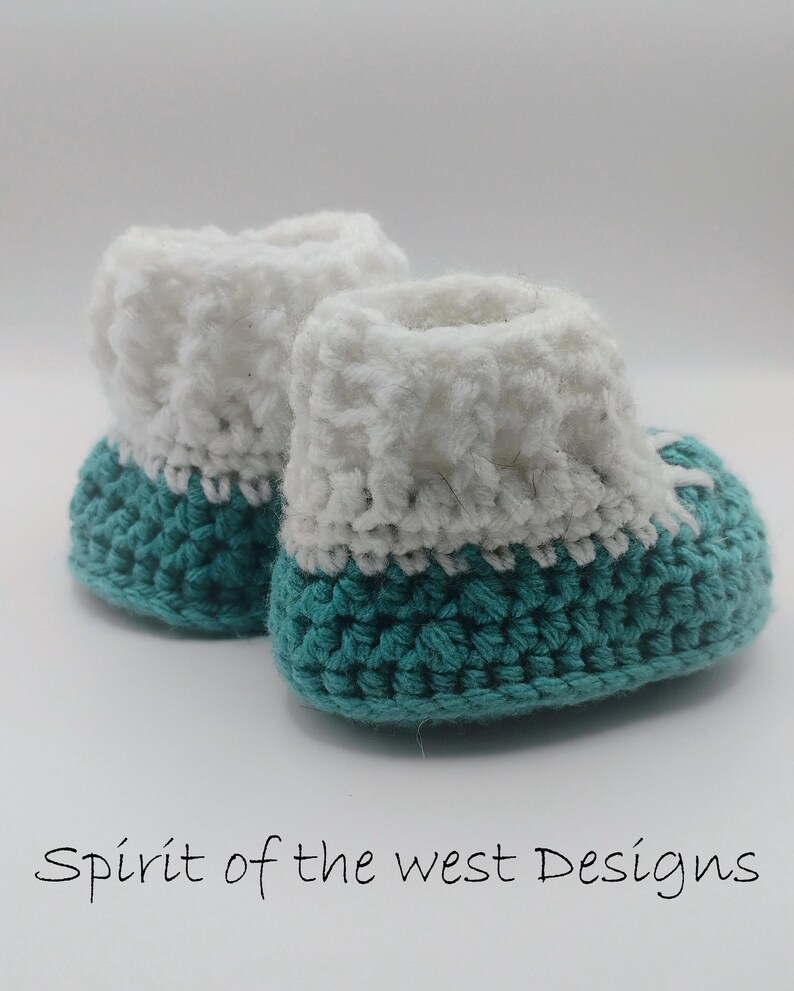 Pu&ograve; includere: Un paio di scarpine da neonato all'uncinetto fatte a mano. Le scarpine hanno una base turchese e un polsino bianco. Il testo "Spirit of the west Designs" &egrave; in basso nell'immagine.