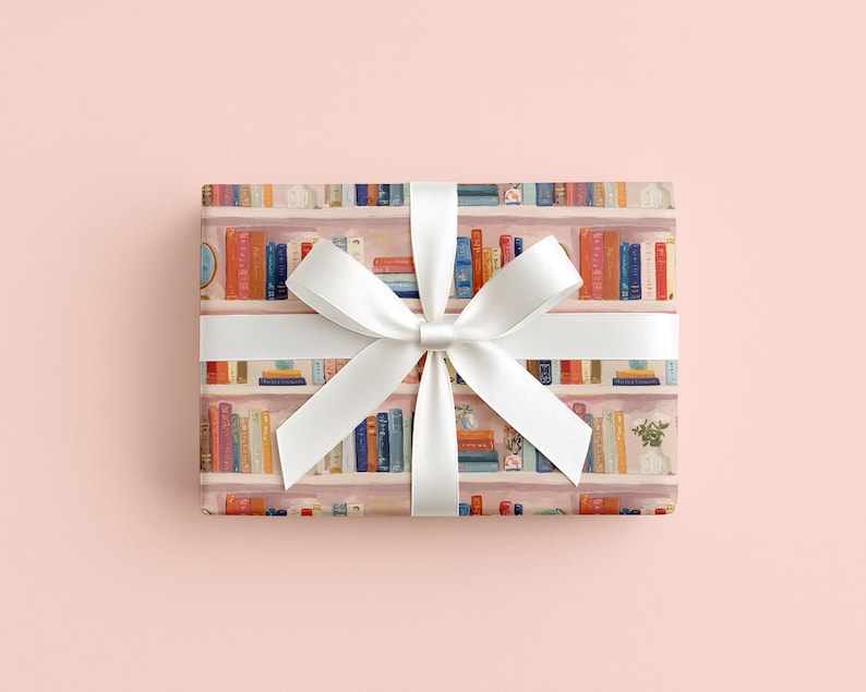May include: A wrapped gift with a white satin ribbon and bow. The wrapping paper features a watercolor design of bookshelves filled with colorful books. The background is a solid light pink.