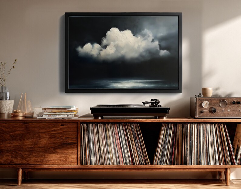 May include: A framed painting of a cloud formation over water, in shades of white, gray, and black, hangs above a wooden cabinet. A black record player sits on top of the cabinet, with a collection of vinyl records stored below.