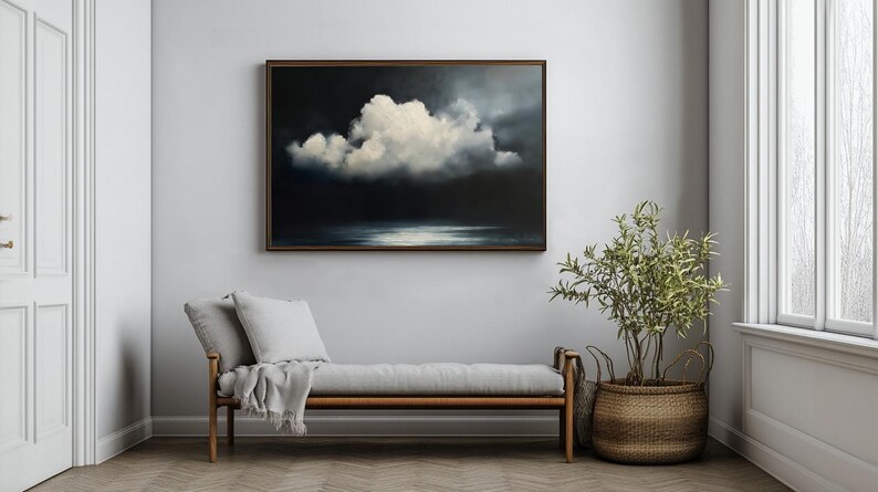 May include: A framed painting of a dramatic cloudscape hangs above a gray bench with pillows and a blanket. A potted plant in a woven basket sits nearby. The artwork features a large white cloud against a dark background.