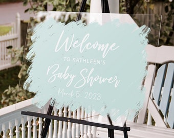 May include: A clear acrylic sign with a light blue brushstroke design. The sign reads "Welcome to Kathleen's Baby Shower March 5, 2023". The sign is on a black easel.