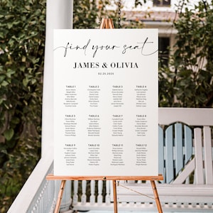 May include: A white wedding seating chart on a wooden easel. The sign reads "find your seat" above the names James & Olivia and the date 02.25.2025. The chart lists guest names under table numbers 1-12.