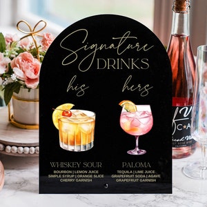 May include: Black sign with gold lettering that reads "Signature Drinks" with a whiskey sour and a paloma cocktail illustrated on either side. The sign also includes the text "his" and "hers" with the drink recipes listed below each cocktail.