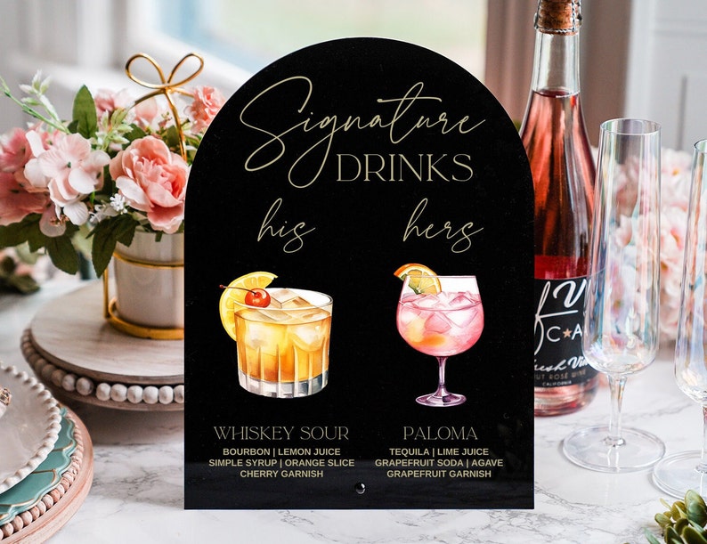 May include: Black sign with gold lettering that reads "Signature Drinks" with a whiskey sour and a paloma cocktail illustrated on either side. The sign also includes the text "his" and "hers" with the drink recipes listed below each cocktail.