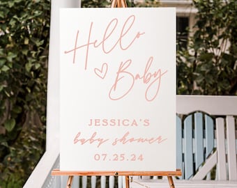May include: A white sign with pink lettering that says "Hello Baby" with a heart symbol. Below the text is "Jessica's Baby Shower" and the date "07.25.24". The sign is on a wooden easel.