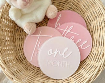May include: A woven basket holds a plush white lamb toy and several pink and white milestone discs. The discs have white script that reads "one month" and other milestone phrases. The discs are in varying shades of pink.