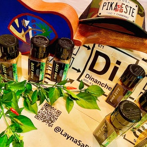 May include: Four glass jars of Pika Este seasoning blend, with a green plant and red peppers in the foreground. The jars have a black lid and a white label with the text "Pika Este" in black. The jars are on a white surface with a black and white QR code and the text "@LaynaSan". A brown ukulele and a camouflage hat with the text "Pika Este" are in the background.