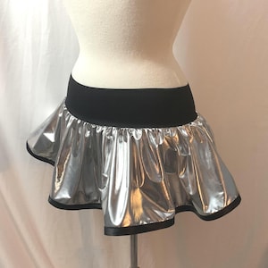 May include: A silver metallic mini skirt with a black elastic waistband and black trim. The skirt has a ruffled, flared design, and is displayed on a mannequin. The shiny material reflects light, creating a dramatic effect.