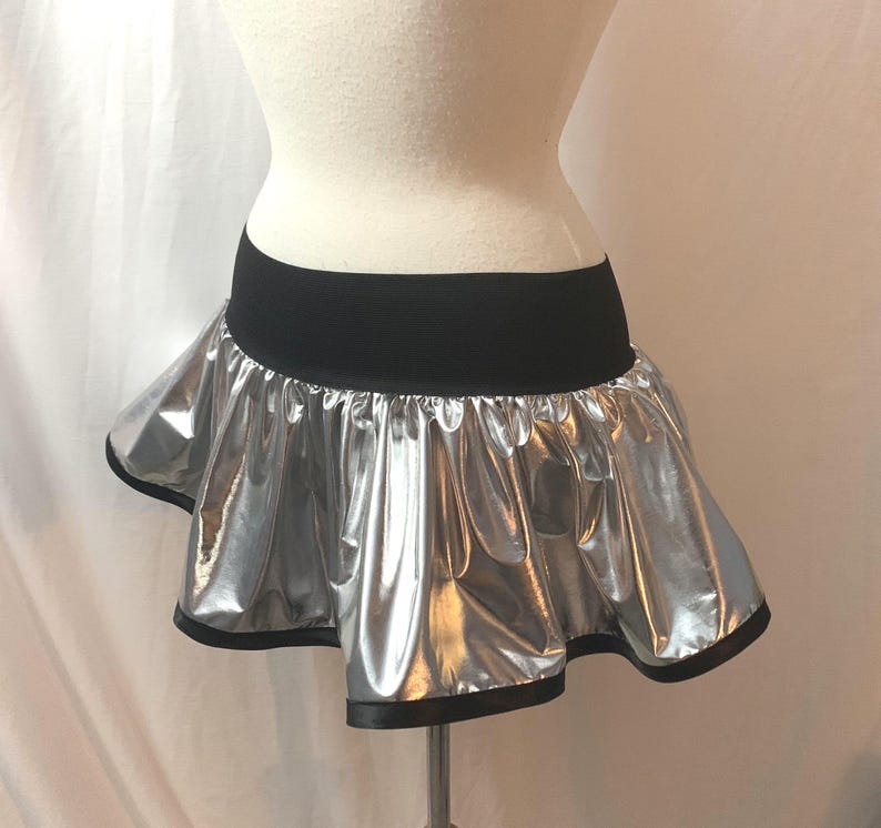 May include: A silver metallic mini skirt with a black elastic waistband and black trim. The skirt has a ruffled, flared design, and is displayed on a mannequin. The shiny material reflects light, creating a dramatic effect.