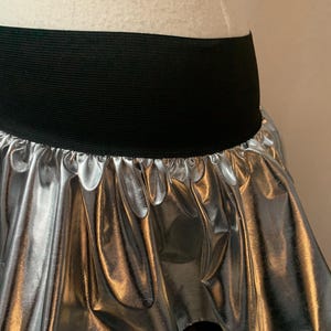 May include: Silver metallic skirt with a black elastic waistband. The skirt has a gathered, ruffled design, creating a flowing, layered effect. The shiny material reflects light, adding a dynamic visual element. This skirt is suitable for dance or costume.
