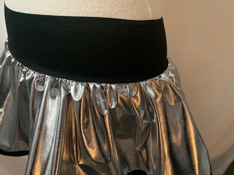 May include: Silver metallic skirt with a black elastic waistband. The skirt has a gathered, ruffled design, creating a flowing, layered effect. The shiny material reflects light, adding a dynamic visual element. This skirt is suitable for dance or costume.