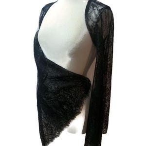 May include: A black lace bolero with long sleeves and an open front design. The garment features a delicate floral pattern throughout the sheer fabric, with scalloped edges along the hem and cuffs, draped over a white mannequin torso.