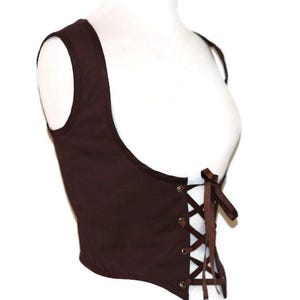 May include: A dark brown sleeveless bodice featuring a wide scoop neckline and a front lace-up closure. The garment is secured with brown ribbon threaded through four pairs of silver-toned metal grommets, tied in a bow at the center.