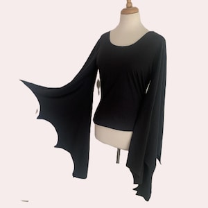 May include: A solid black long-sleeve top featuring a scoop neckline and wide, flared sleeves with a scalloped, bat-wing edge design. The garment is displayed on a mannequin, showcasing the flowing fabric and the distinct pointed hemline of the sleeves.