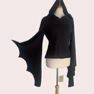 May include: A black long-sleeved top featuring an attached hood and wide, flared sleeves with a scalloped, bat-wing edge. The garment is displayed on a mannequin, showcasing the solid black fabric and the distinctively shaped sleeve hems.