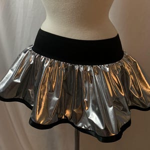 May include: A silver metallic mini skirt with a black waistband and trim. The skirt has a ruffled, bubble-like design, creating a voluminous silhouette. The skirt is displayed on a mannequin.