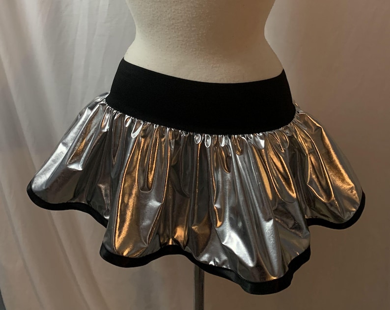 May include: A silver metallic mini skirt with a black waistband and trim. The skirt has a ruffled, bubble-like design, creating a voluminous silhouette. The skirt is displayed on a mannequin.