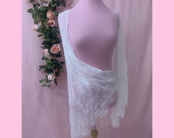 Peut inclure: Un châle en dentelle blanche drapé sur un mannequin. Le tissu présente un motif floral délicat et complexe avec des bords festonnés. La matière légère est semi-transparente, laissant passer la lumière à travers le tissage fin.