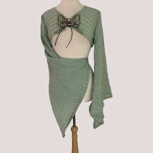 May include: A light sage green long-sleeved wrap top featuring a textured fabric. The garment includes a decorative butterfly-shaped patch at the center chest with dark brown lace-up detailing and long, flowing sleeves that drape down the sides.