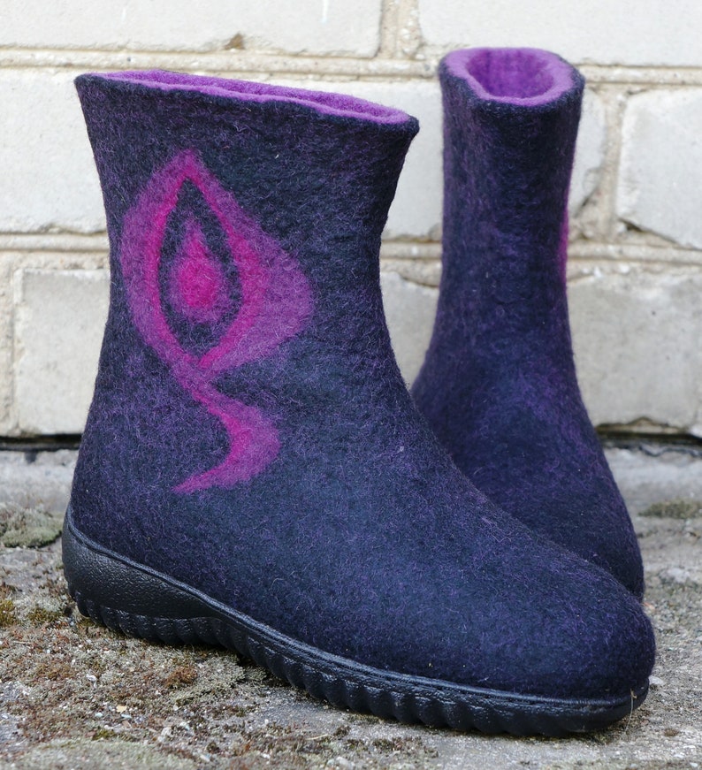 May include: A pair of dark purple felt boots with a pink design on the front. The boots have a black rubber sole.