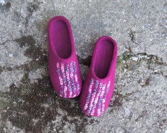 May include: A pair of magenta felt slippers. Each slipper has a decorative pattern of vertical lines in shades of white, gray, and pink. The slippers are shown on a concrete surface.