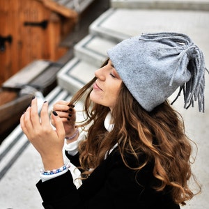 May include: A woman wearing a grey wool hat with long fringe and a black jacket. She is sitting on a set of concrete steps.