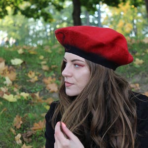 Op de afbeelding: Een vrouw met lang bruin haar draagt een rood-zwarte baret. Ze kijkt naar de zijkant en heeft een bedachtzame uitdrukking op haar gezicht.