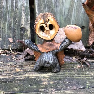May include: A handcrafted woodland creature figurine made from natural materials. It features a walnut shell head, a leaf cape, and an acorn held in one hand. The body is made of a textured, grey material.