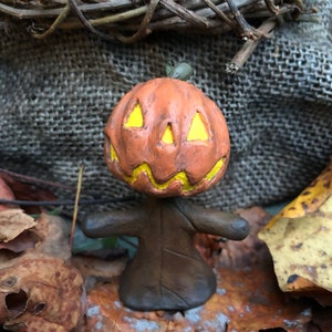 May include: A small, handmade pumpkin figurine with a carved face and yellow paint accents. The figurine is standing on a bed of brown leaves.