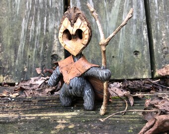 May include: A whimsical, handmade woodland creature figurine. The figure is crafted from a walnut shell, with a heart-shaped face, and wears a leaf cape and a brown leather vest. It holds a small twig, standing on a wooden surface.