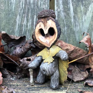 Può includere: Figurina artigianale di una creatura del bosco, realizzata con un guscio di noce. La figura ha un volto scolpito, un cappello di ghianda e un mantello di foglie gialle. Tiene un piccolo bastone, tra foglie secche.