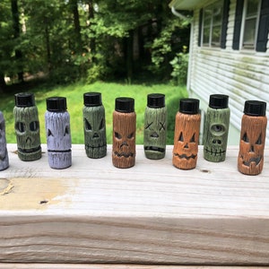 Può includere: Una collezione di dieci piccole bottiglie decorative con tappi neri. Ogni bottiglia è scolpita con un design unico di volto stilizzato in vari colori, tra cui grigio, verde e marrone. Le bottiglie sono disposte su una superficie di legno, suggerendo un tema di Halloween o novità. Circa 10 cm di altezza.