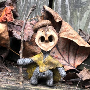 May include: A whimsical, handmade woodland creature figurine. The figure is crafted from a walnut shell, acorn, and other natural materials. It has a gray body, a yellow leaf cape, and holds a small stick. The background features dried leaves.