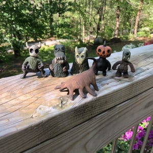 Puede incluir: Cinco pequeñas figuritas de cerámica pintadas a mano, que incluyen una calabaza, una criatura con un hocico largo, una criatura con una máscara, una criatura con cuernos y una criatura con un cuerpo largo y delgado. Las figuritas están dispuestas sobre una superficie de madera.