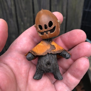 May include: A small, handcrafted figurine made from an acorn shell. The acorn shell is carved into a jack-o'-lantern face and is attached to a small, furry body with a brown, orange, and black cloak.