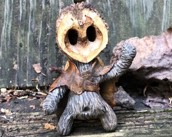 May include: A whimsical, handmade figurine crafted from a walnut shell, featuring a face with large eye sockets. The figure has a brown leaf cape and textured gray arms and body, giving it a rustic, woodland aesthetic.